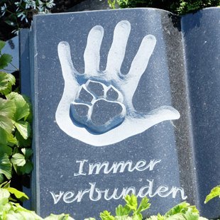 Dunkler Grabstein in Form eines aufgeklappten Buches mit Symbol aus Tierpfote und Hand eines Menschen